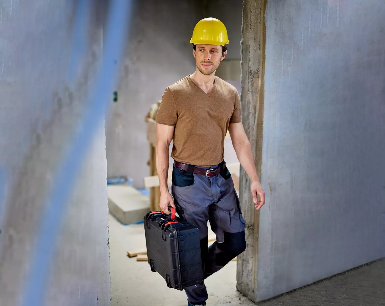 A person in a yellow hard hat carrying a black toolbox in a construction site.