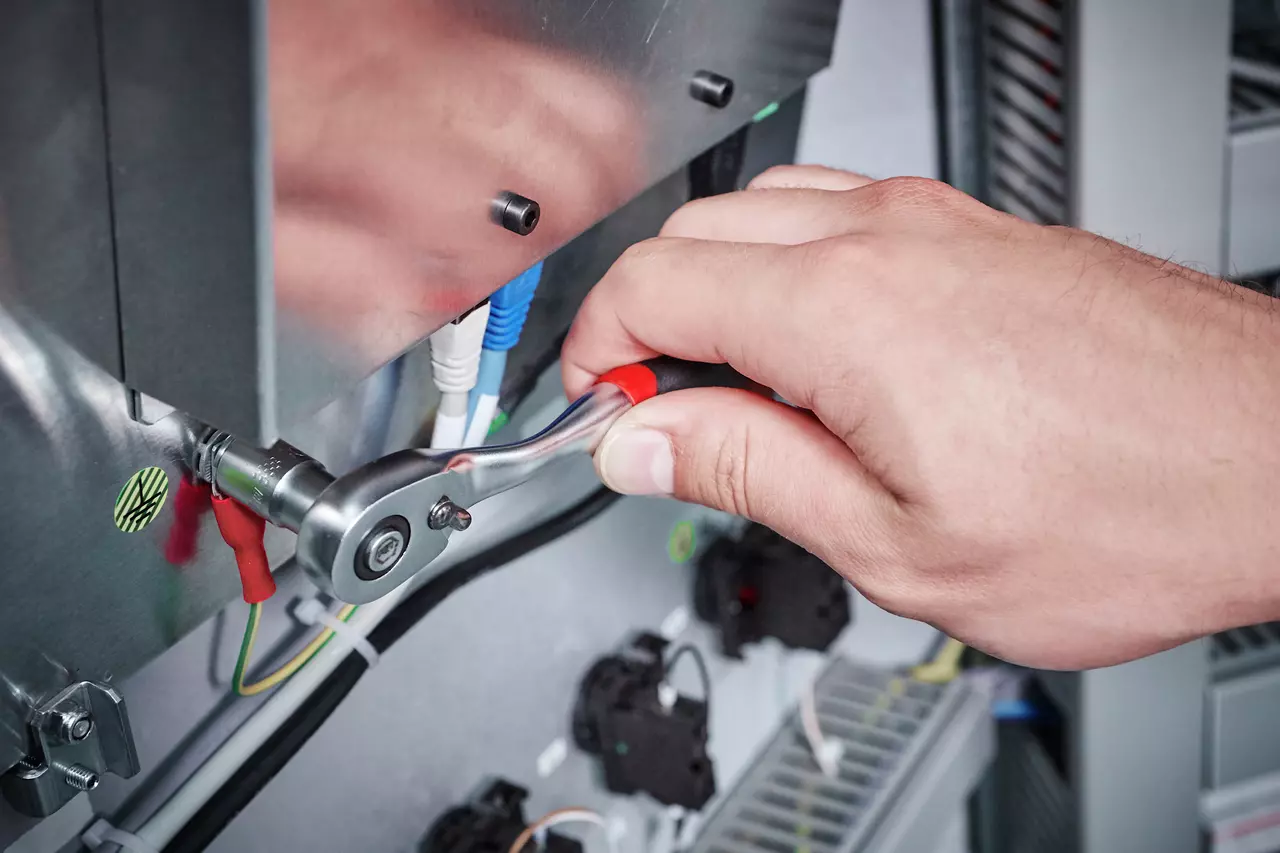 A hand using a ratchet wrench on a metal panel in an electrical setup.
