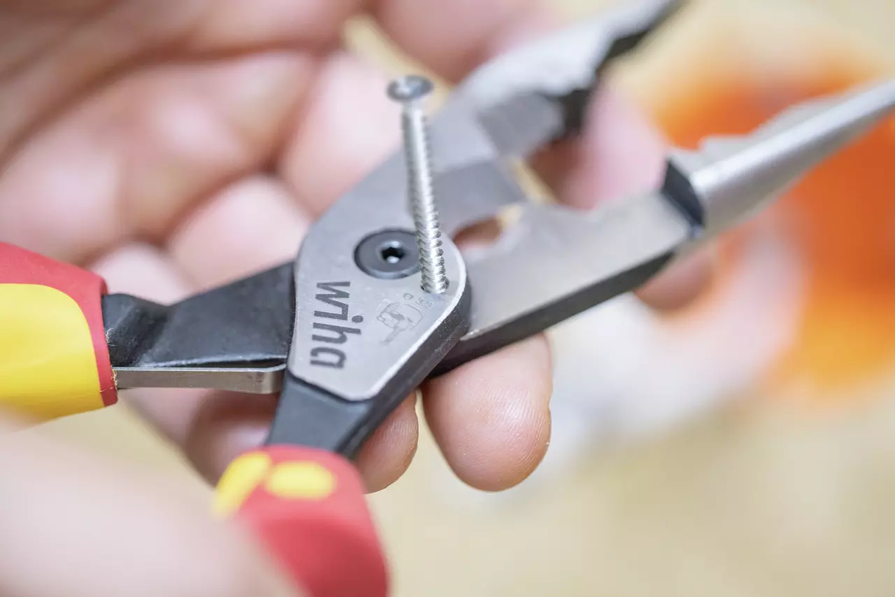 Een hand houdt combinatietang met rode en gele handvatten vast die een schroef grijpt.