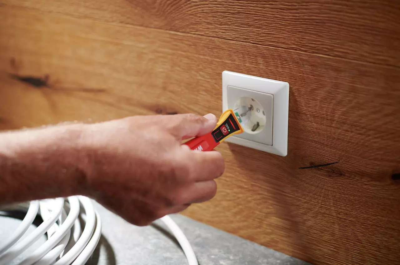 A person uses a non-contact voltage detector near an electrical outlet on a wooden wall.
