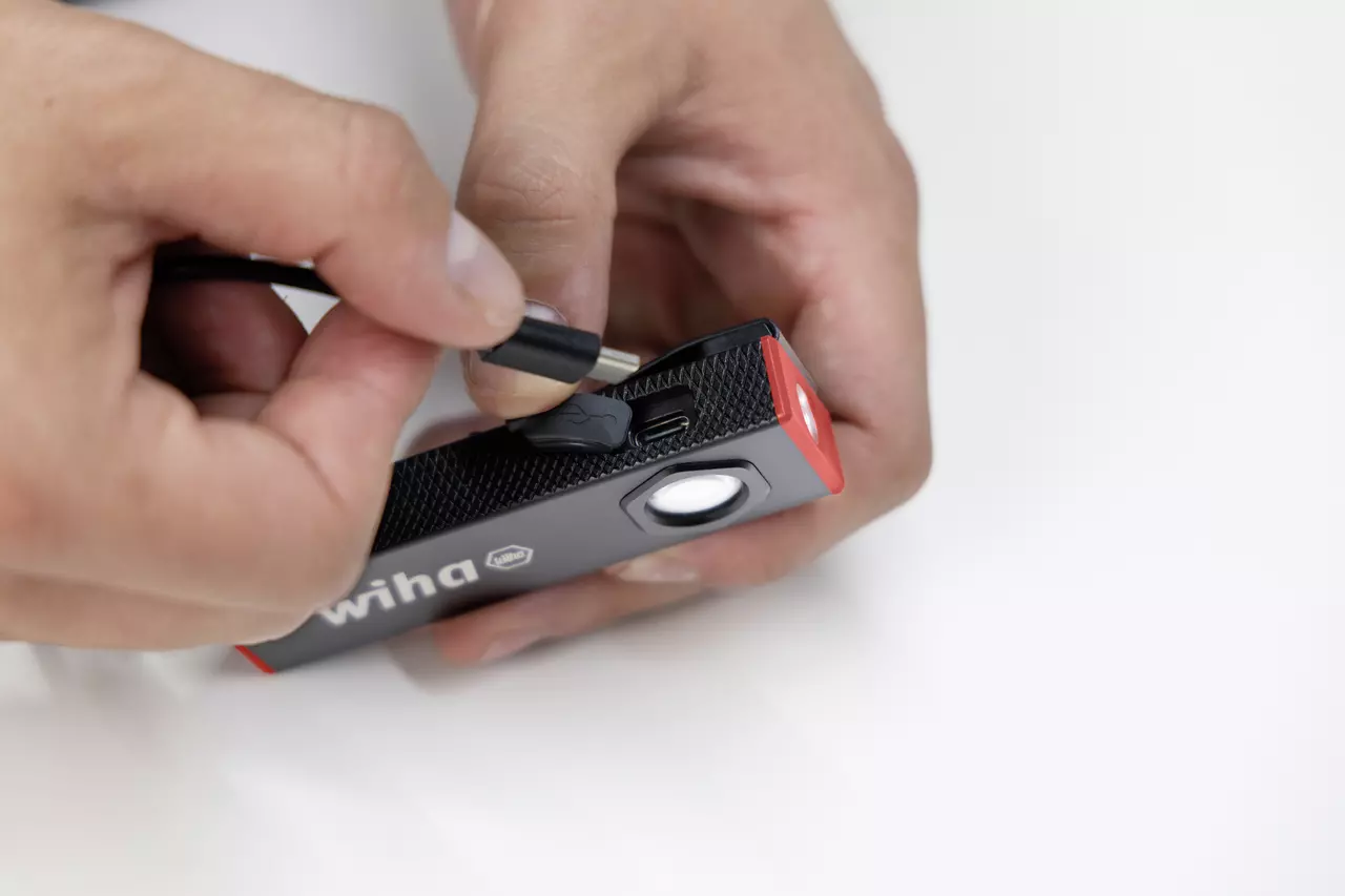 Hands plugging a cable into a black and red flashlight on a white surface.