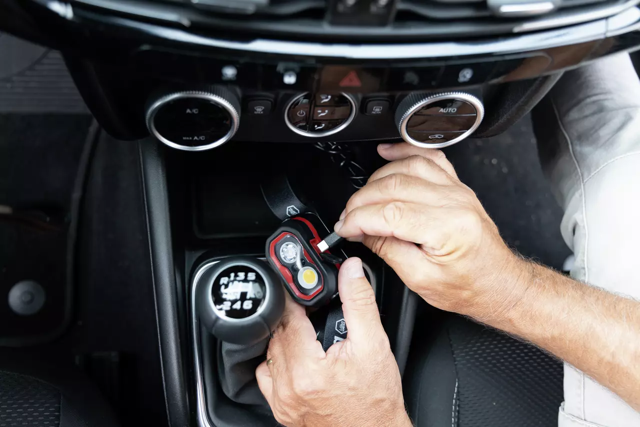 Hands plugging in a cable to charge a headlamp inside a car near the gear shift.