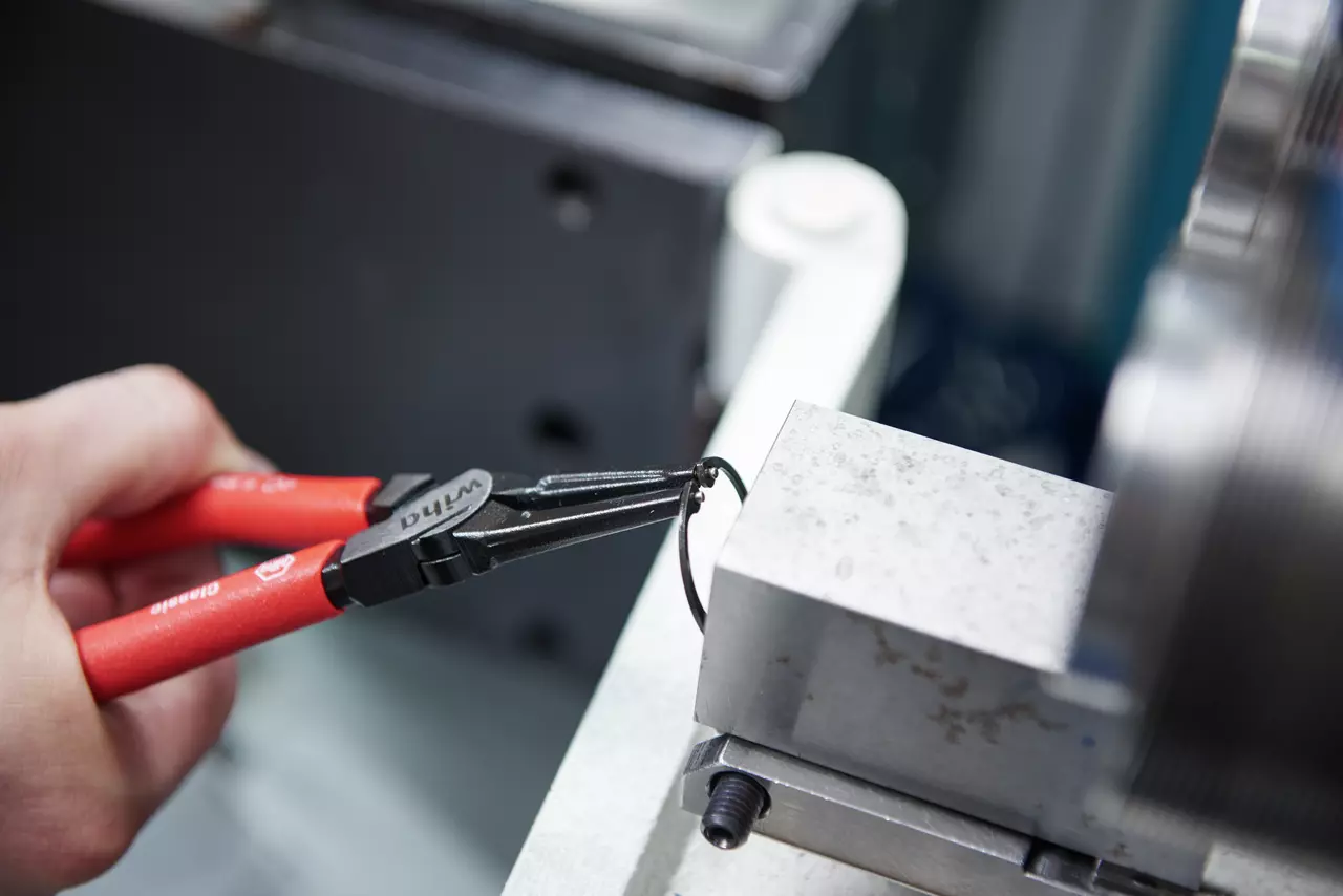 A person uses pliers to remove a retaining ring from a metal block.