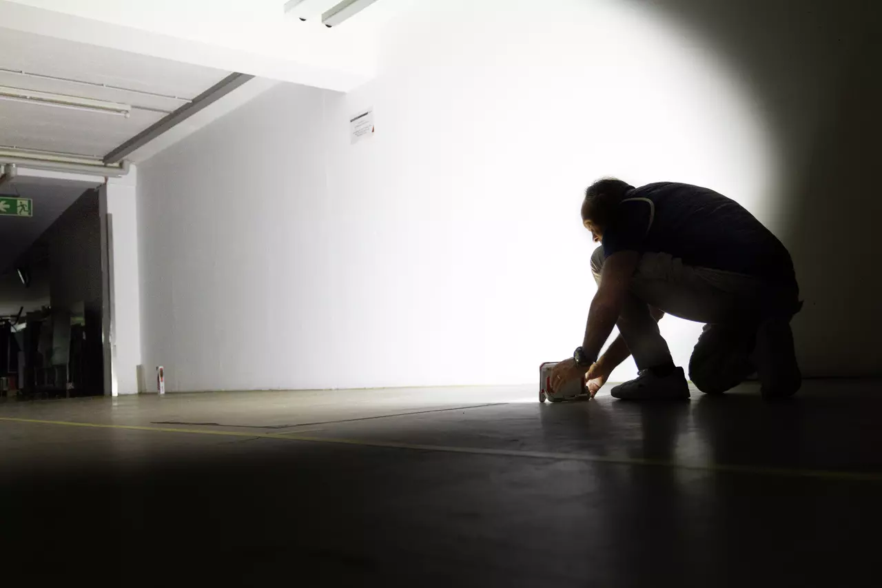 A person kneels in a dark room, holding a square device that projects light onto the wall.