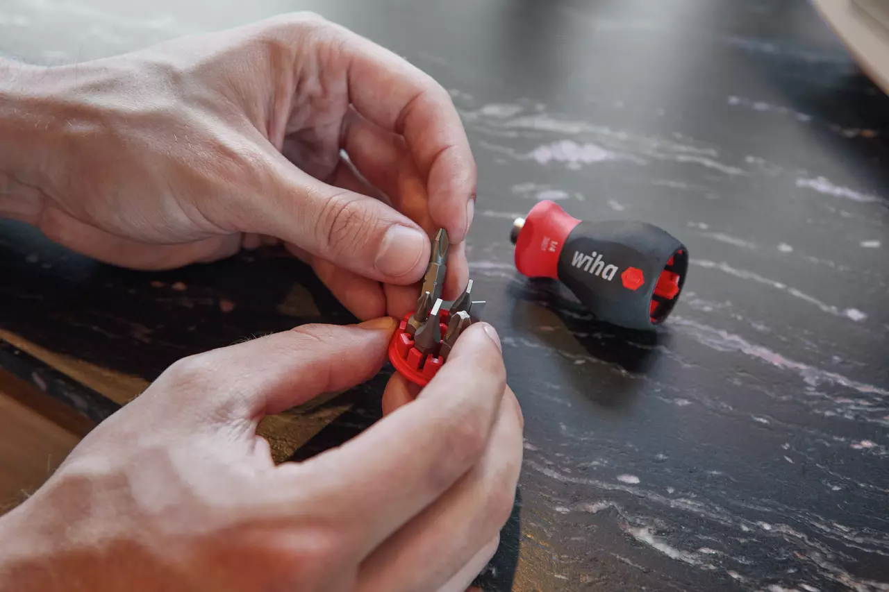 Hands holding a red bit holder with screwdriver bits, next to a black and red screwdriver handle on a table.