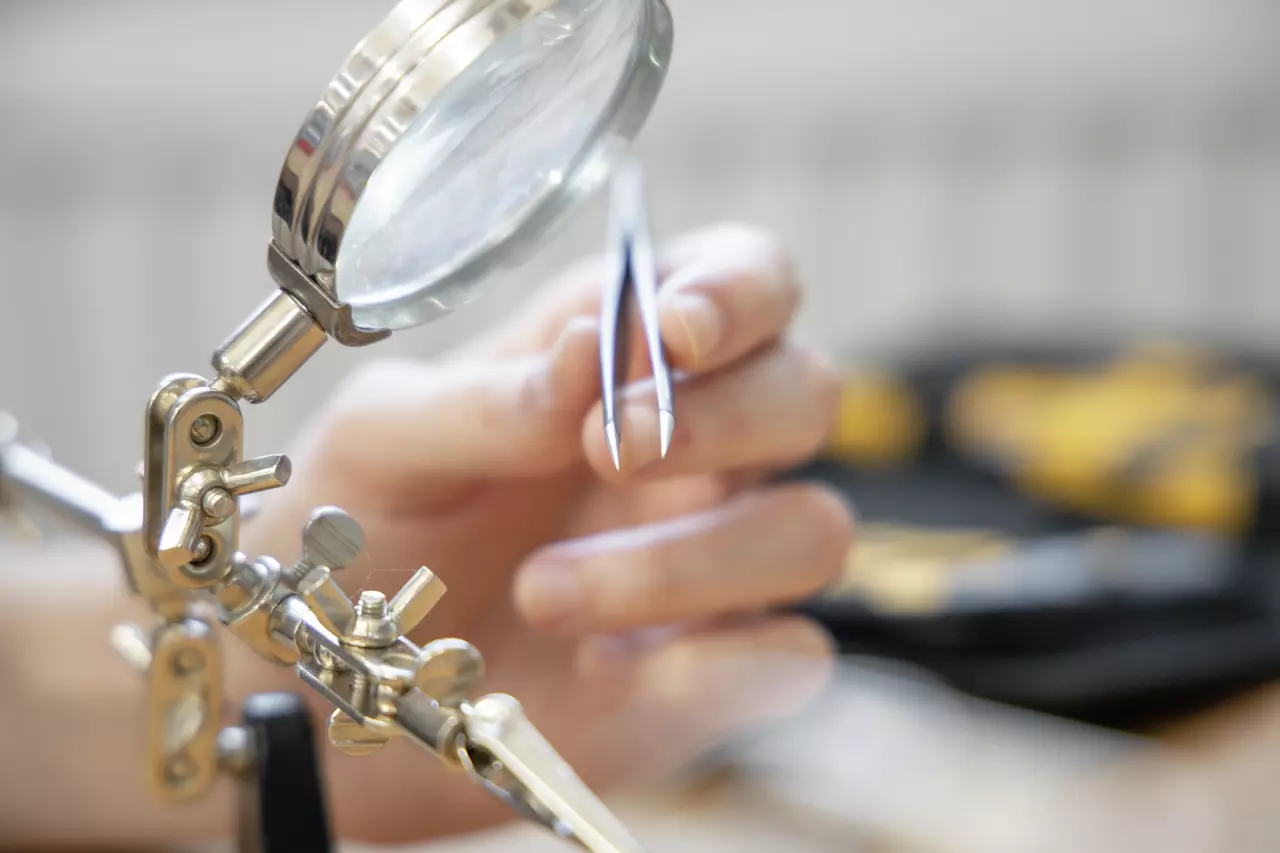 A hand holds tweezers near a magnifying glass on a stand, used for precision work.