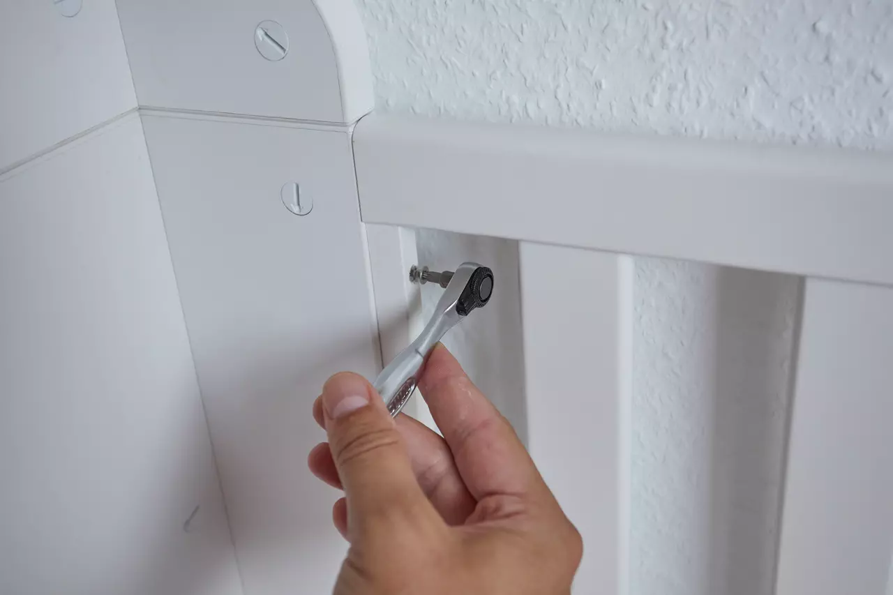 A hand uses a ratchet wrench to tighten a screw on a white piece of furniture.