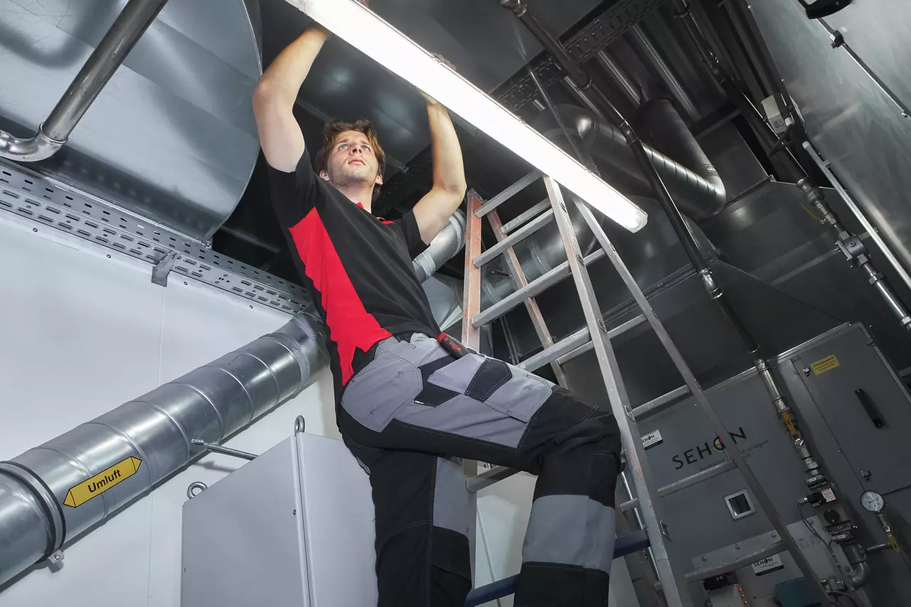 Person working on a ceiling light in a technical room with ventilation ducts.