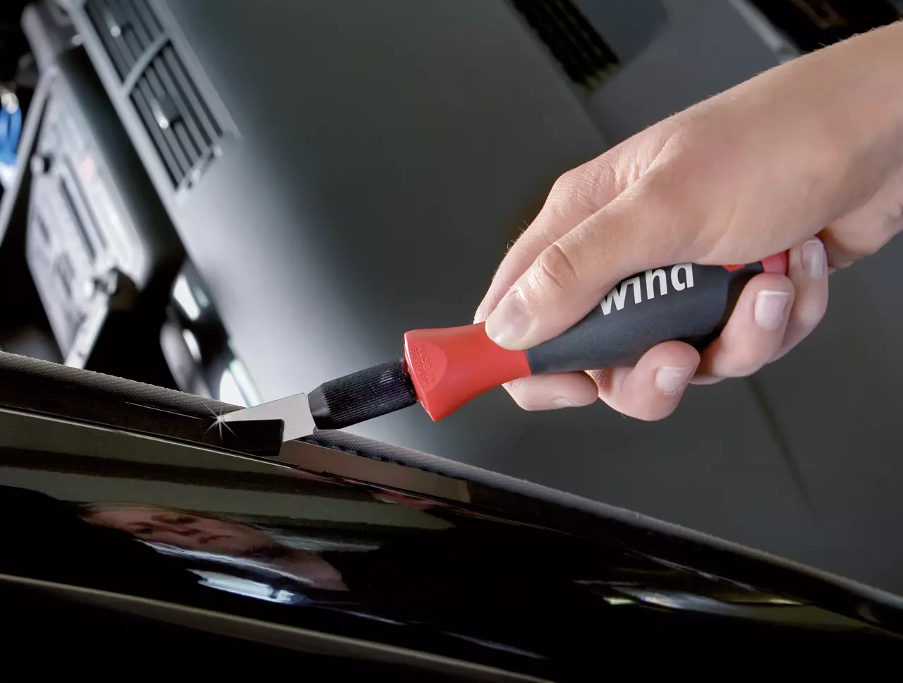A person uses a red and black scraper tool on a car surface.