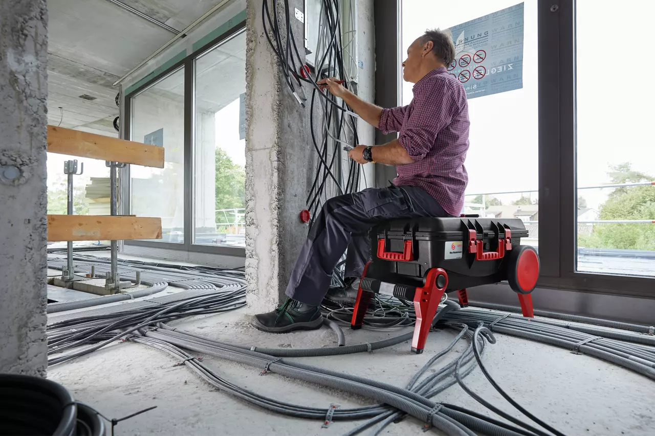 Person sitting on a black tool case, working on cables in a building under construction.
