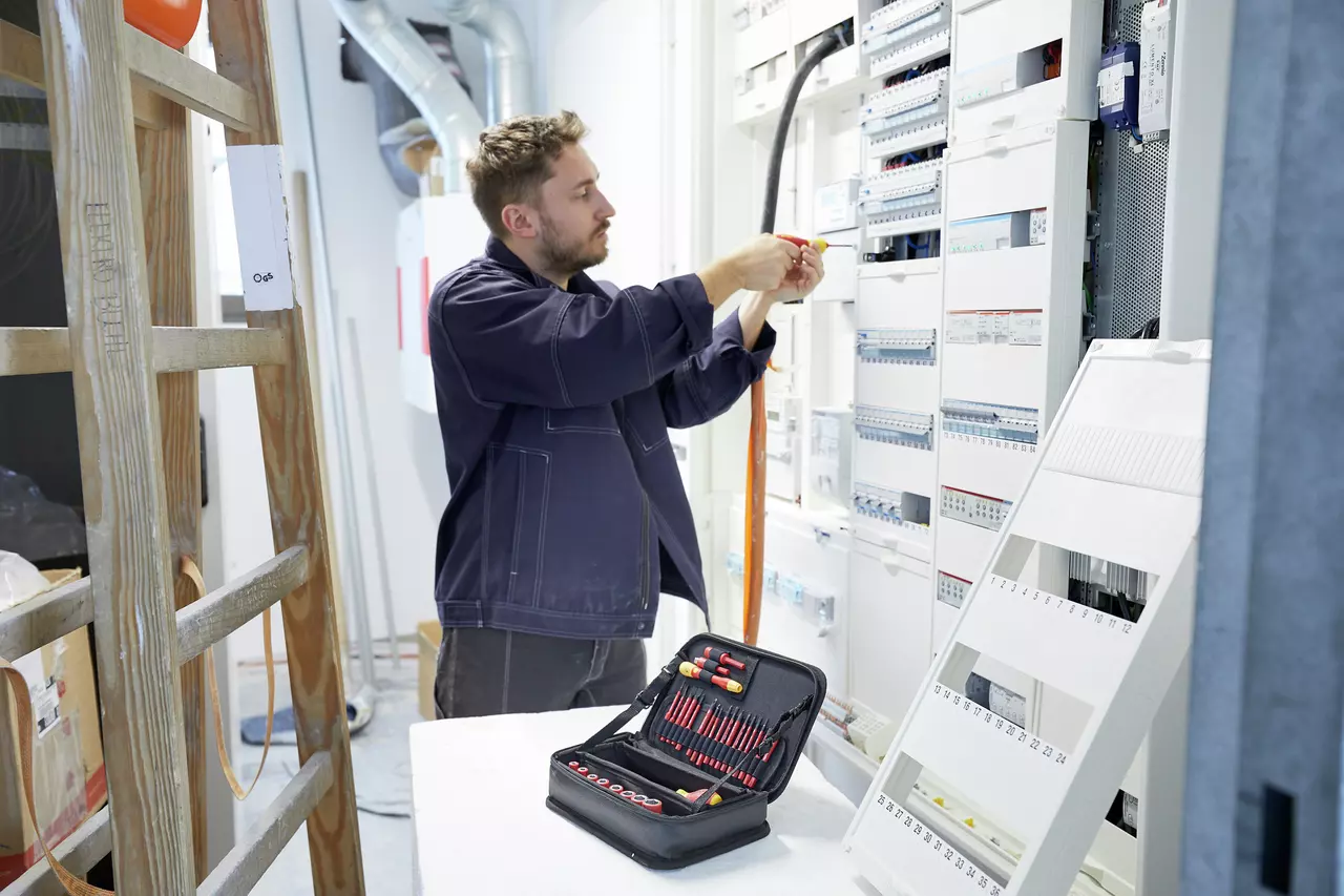A person working on an electrical panel with tools in a case nearby.