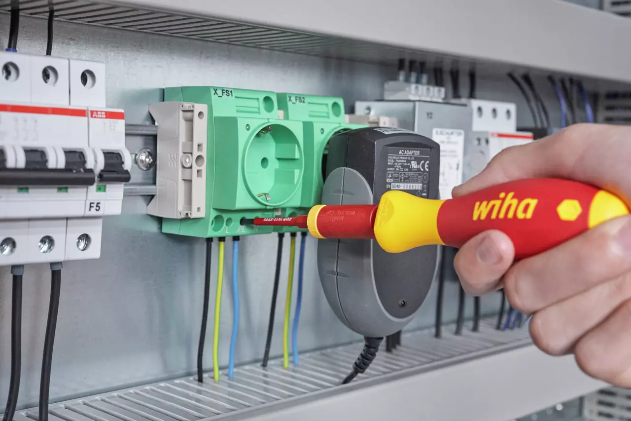 A person uses a red and yellow insulated screwdriver on an electrical panel with green sockets.